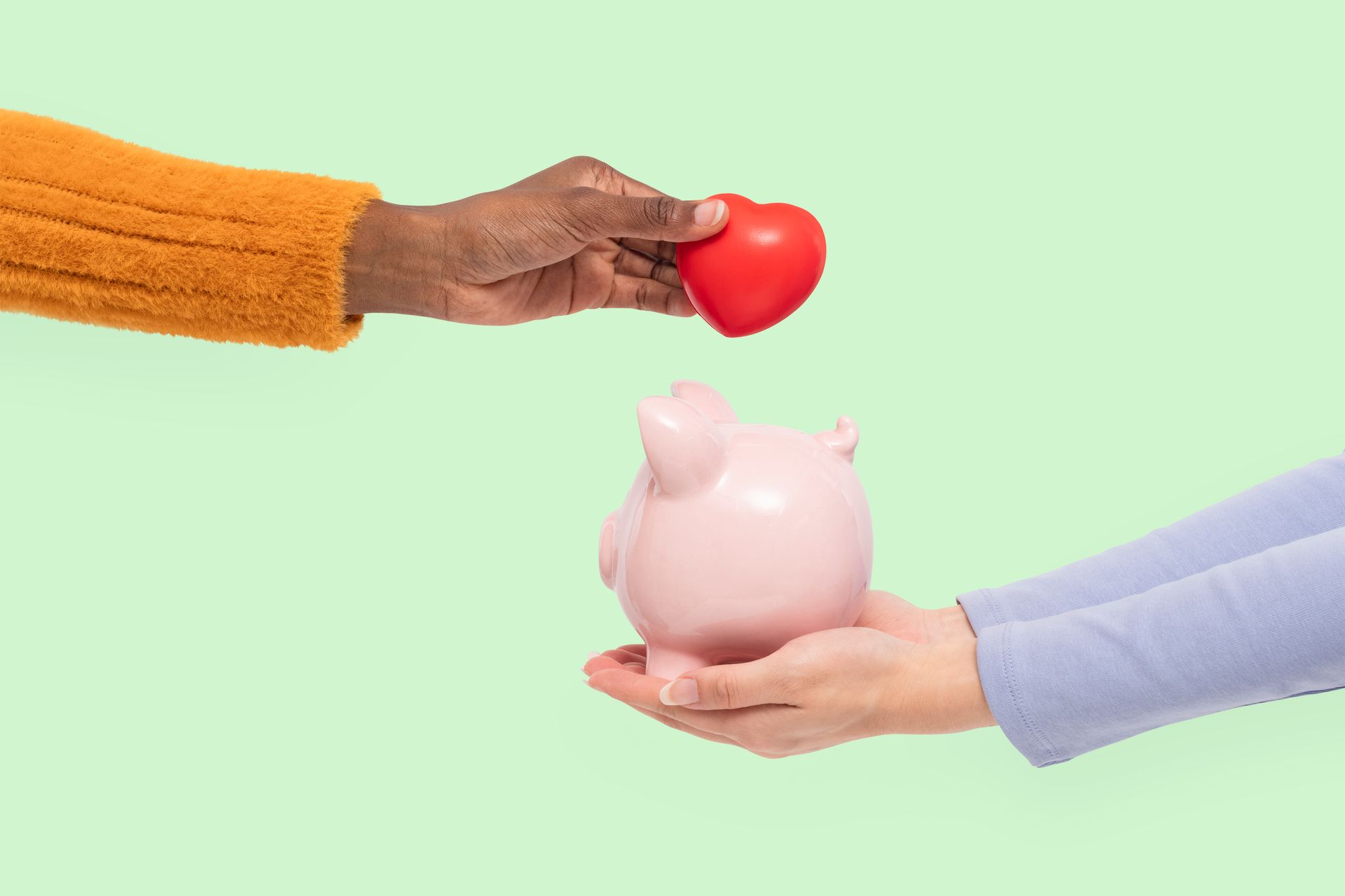 Two hands exchange a red heart over a pink piggy bank on a light green background