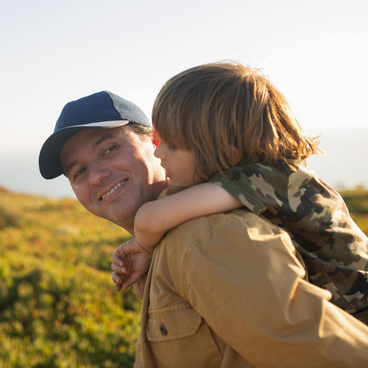 Smiling adult in a blue cap carrying a child in a camo shirt outdoors at sunset