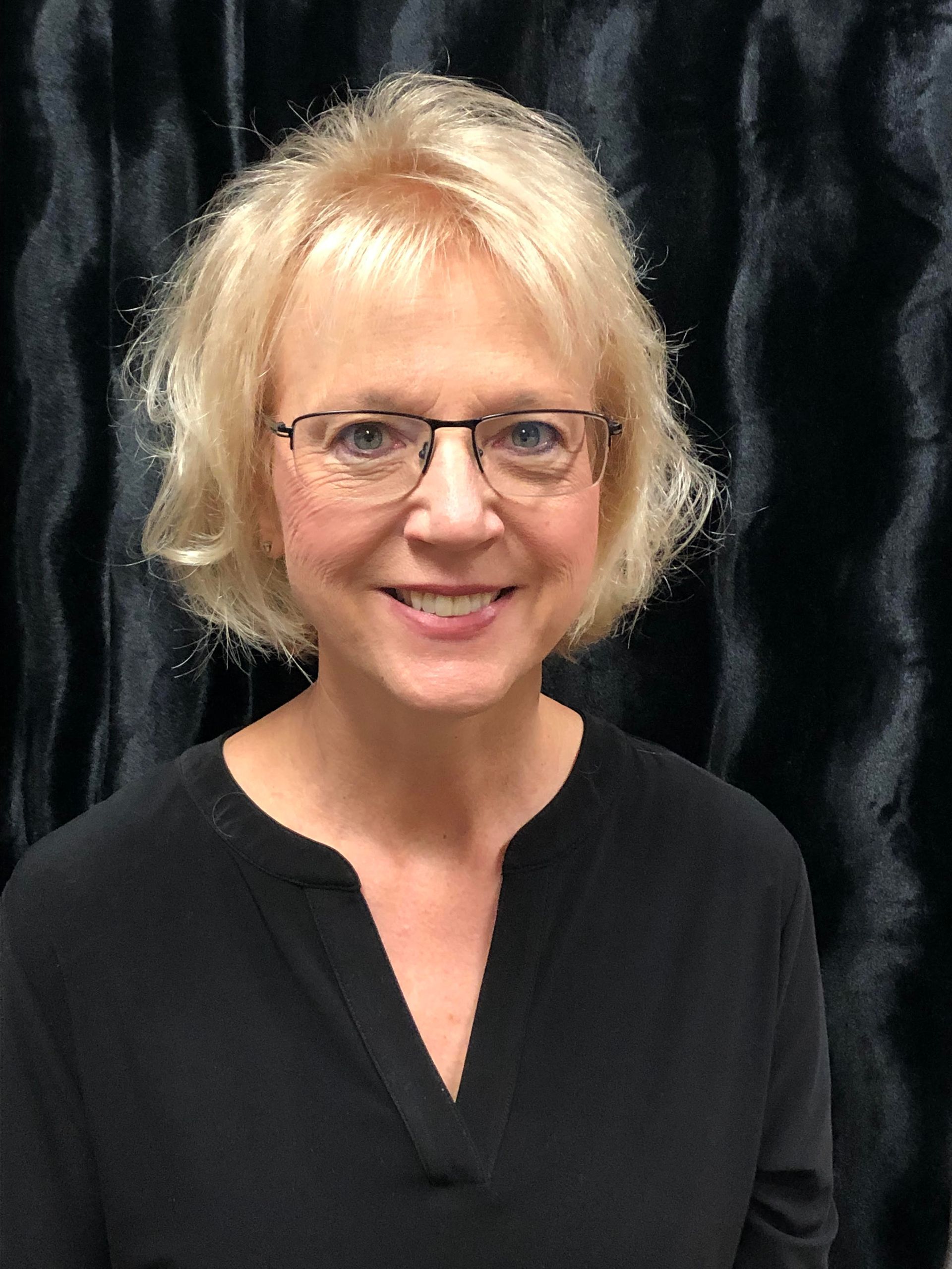 Portrait of a smiling woman with blonde hair and glasses against a dark backdrop