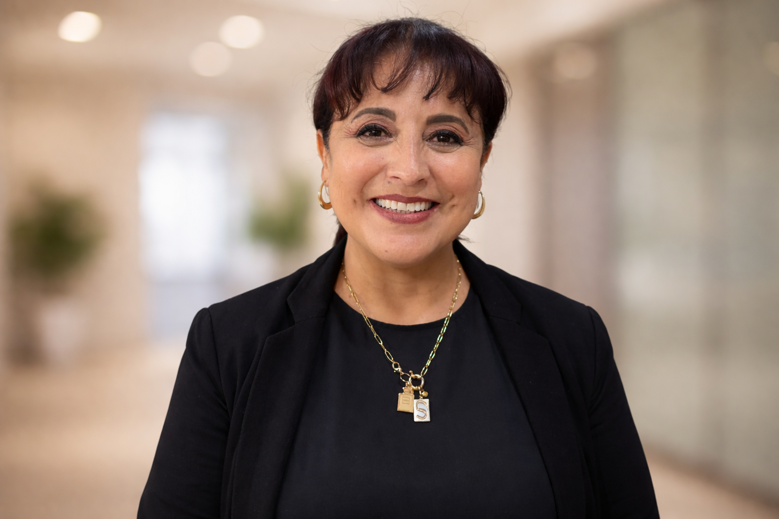 Smiling woman in a black blazer standing in a blurred hallway, wearing gold necklaces.