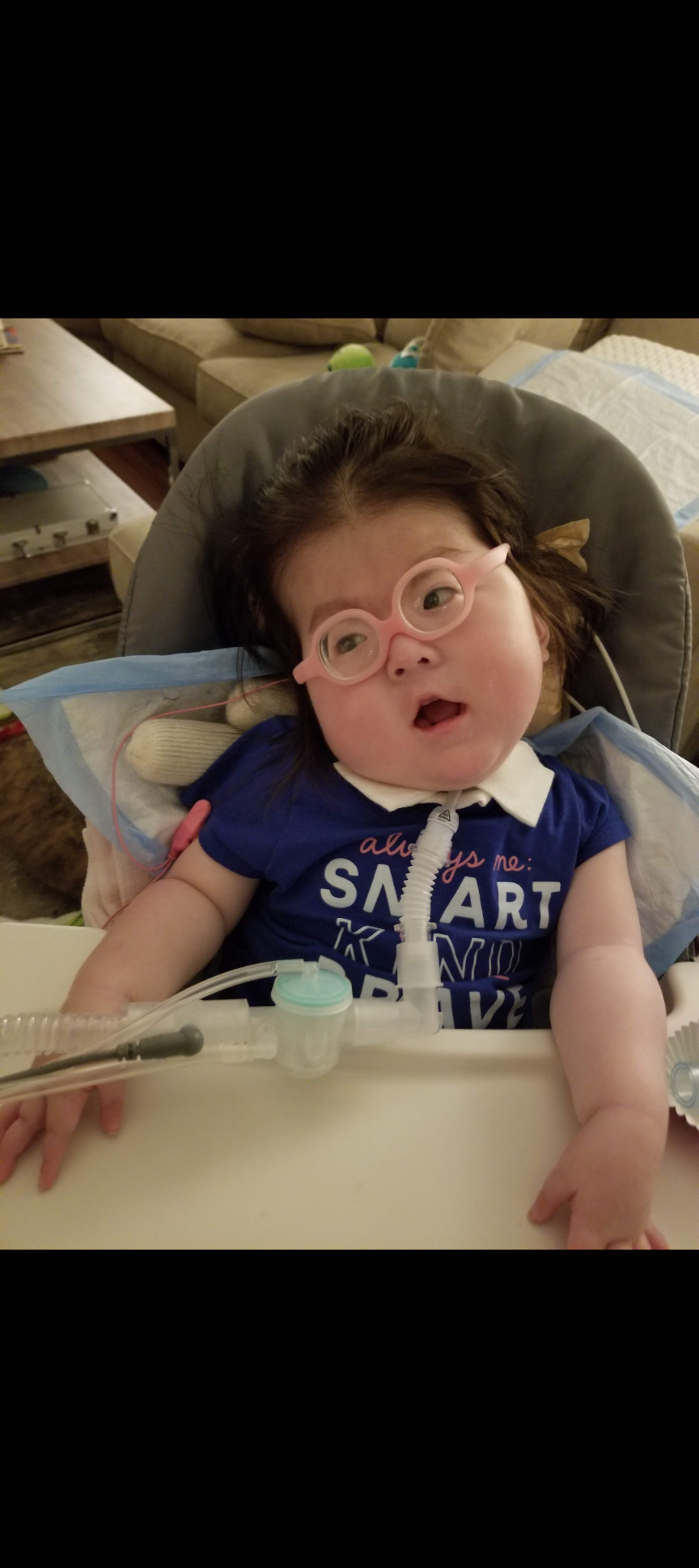 Child with glasses and blue shirt sitting in a stroller, leaning back with mouth open in a playful expression.