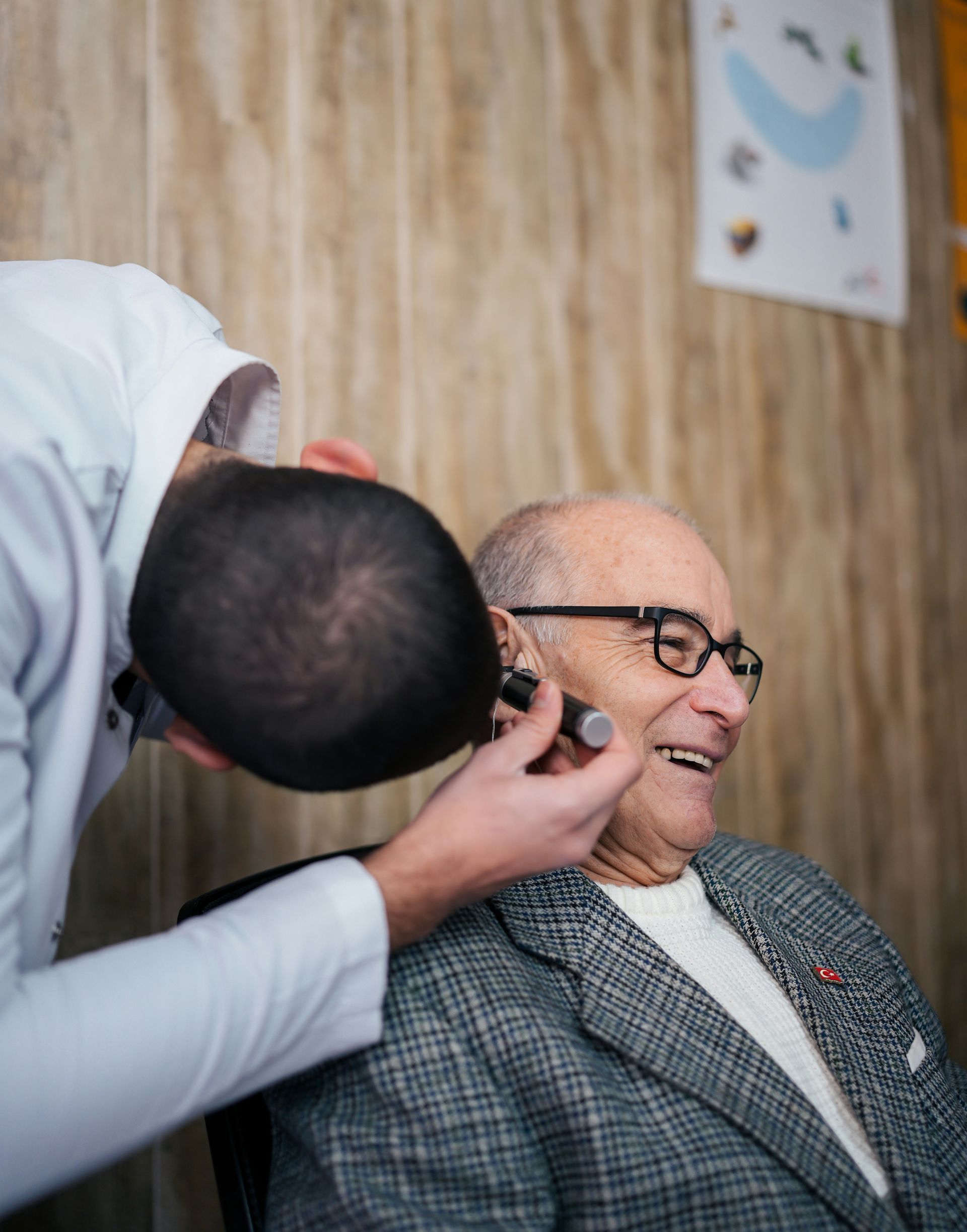 Man trimming an elderly man's beard in a barber chair, with a wood-paneled wall behind them