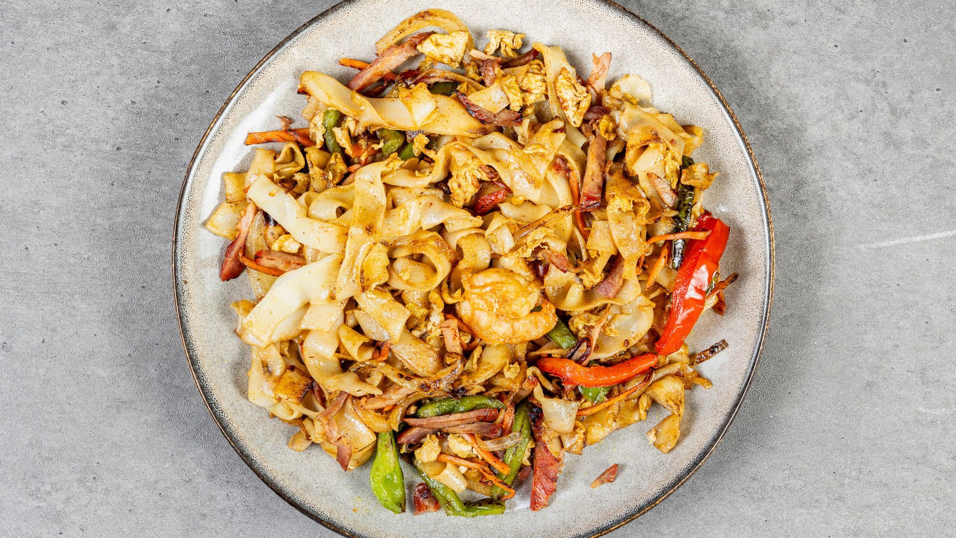 Plate of stir-fried noodles with vegetables and red peppers on a gray surface
