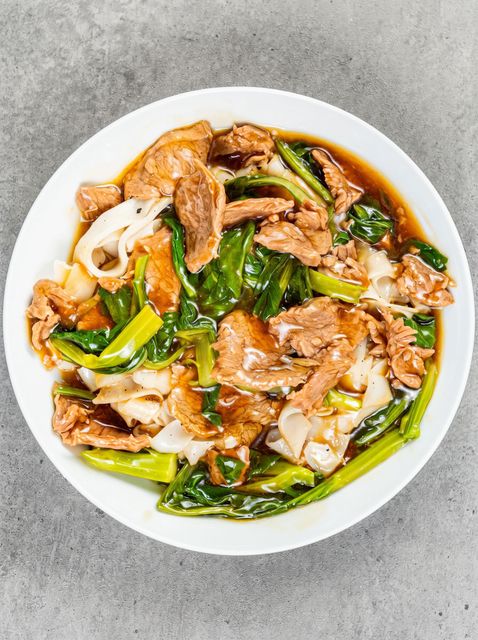 Stir-fried noodles with beef and green vegetables in a white bowl on a gray surface