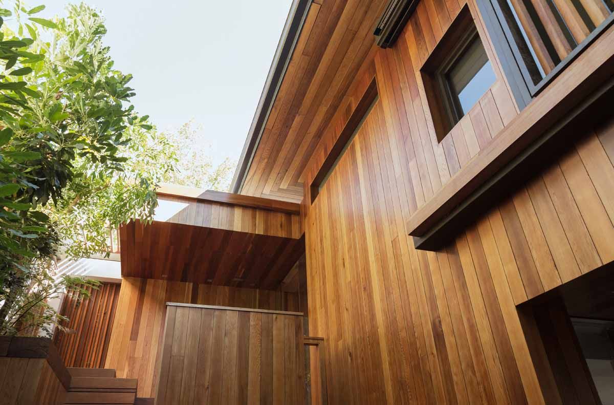 Wooden house exterior with warm brown siding, stairs, and a small window. Lush green tree partially obscures the upper left.