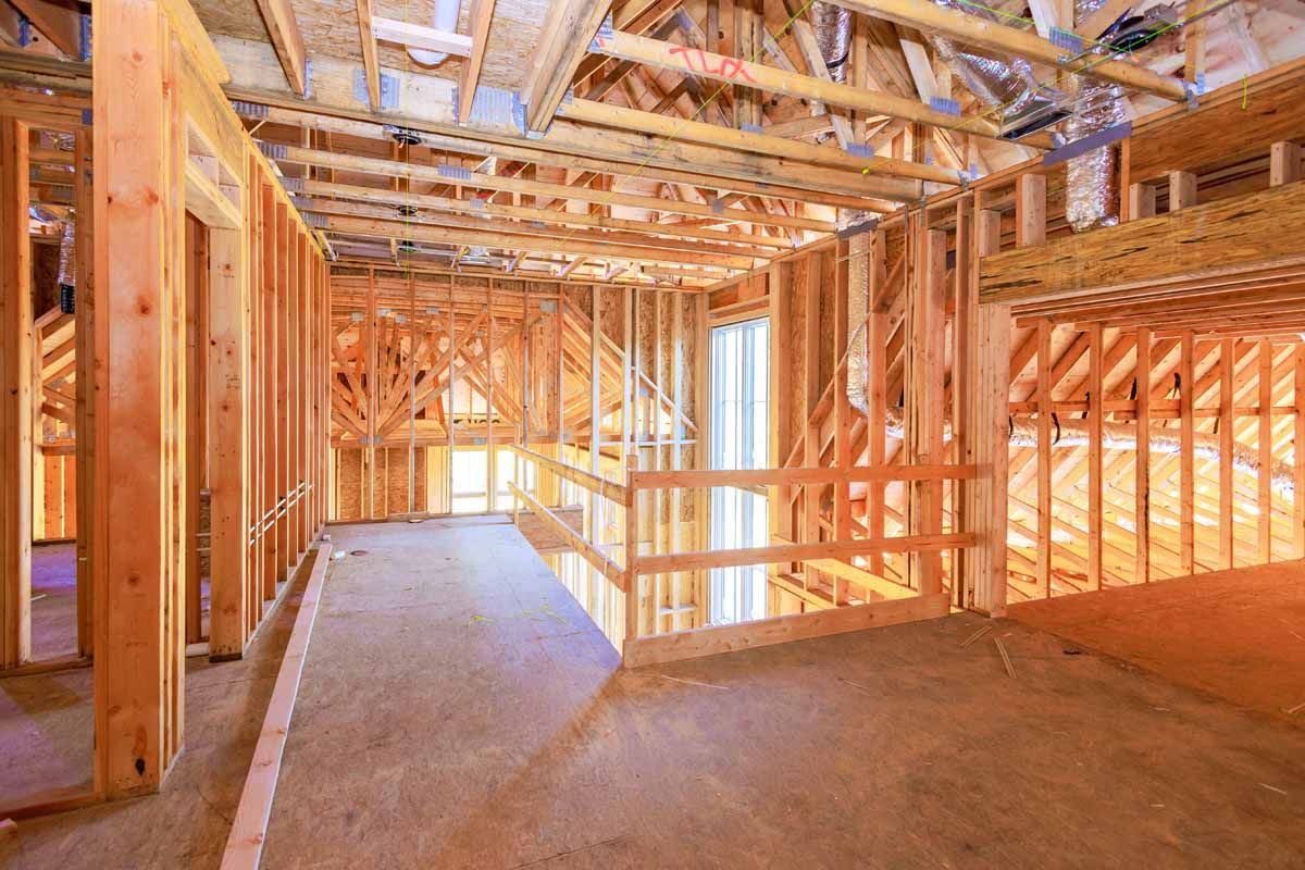Interior view of a house under construction, showing exposed wooden framing for walls, floors, and a balcony. Natural light streams in.