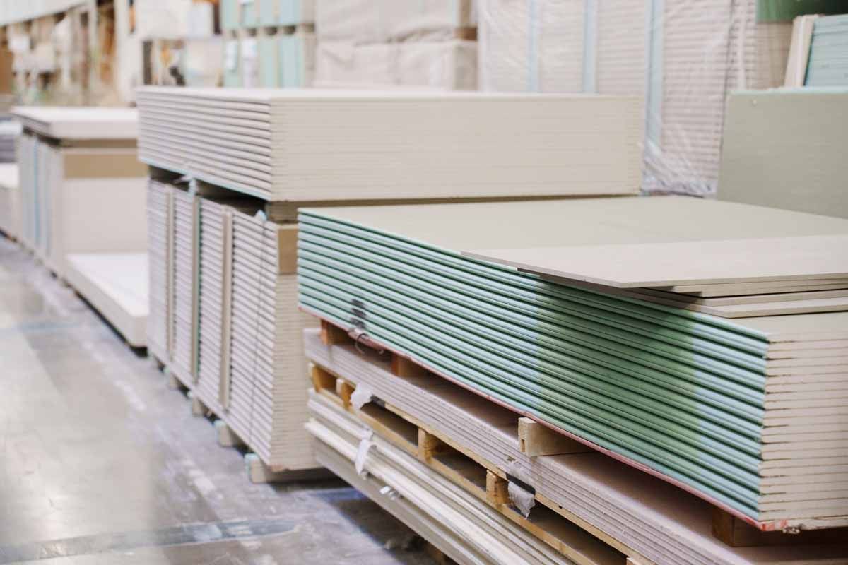 Stacks of drywall boards on pallets, inside a warehouse setting. The boards vary in color, with green and beige visible.