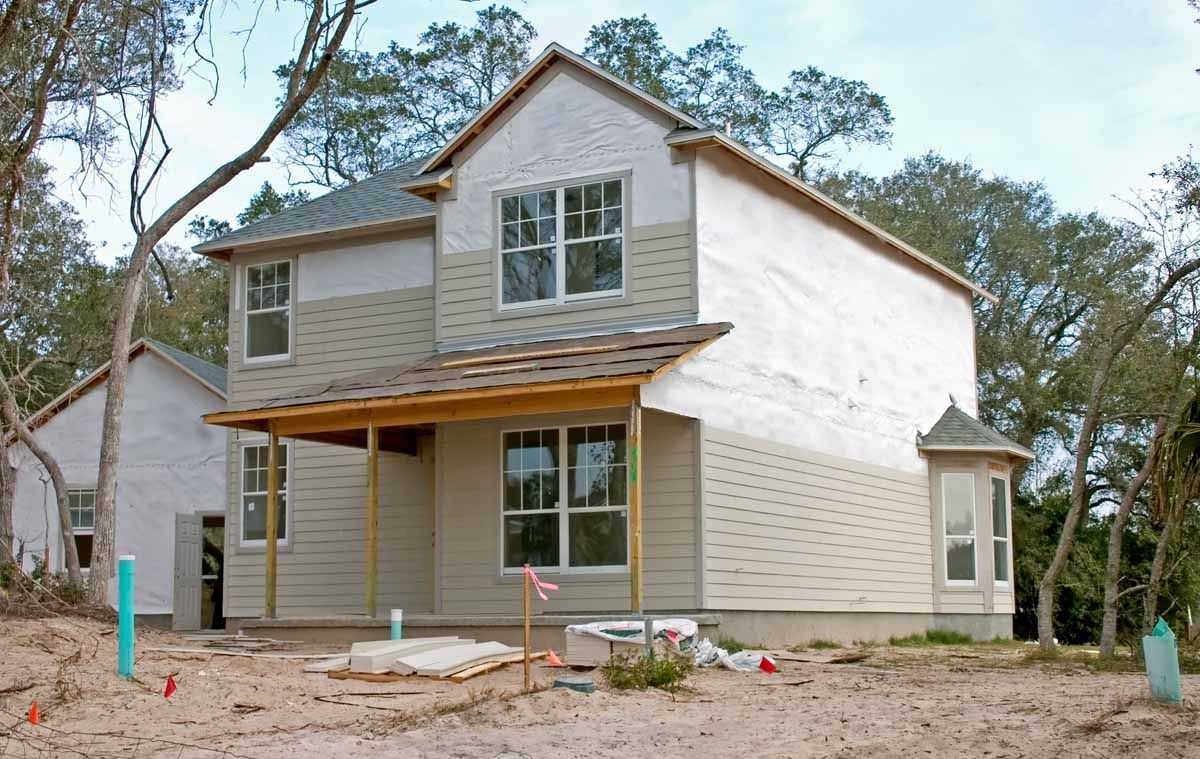 Two-story house under construction in a wooded area; exterior walls are partially covered with beige siding and white paper.