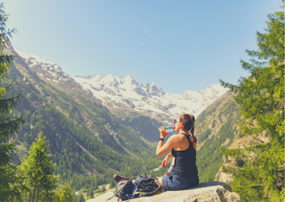 A woman is drinking water from a bottle while sitting on a rock in the mountains.