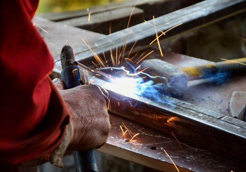 Person welding metal frame; sparks fly, wearing gloves, outdoors.