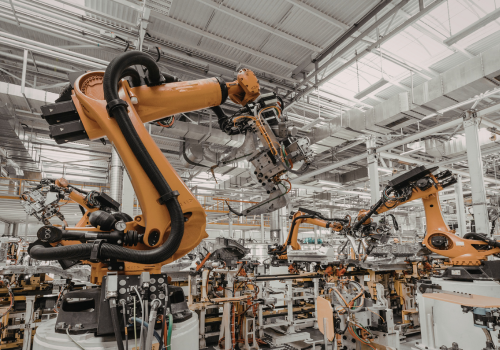 Bright yellow industrial robotic arms operating on a vehicle assembly line in a large, brightly lit factory.