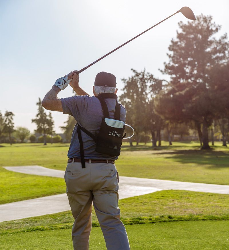Golfer swinging club on a golf course, with a black backpack. Green grass, trees, and sunlight visible.