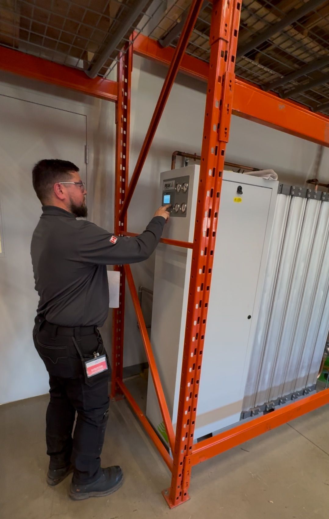 Man in gray uniform operating a machine panel within orange industrial shelving.