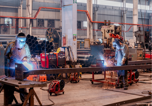 Two welders working on metal beams in a large industrial facility.