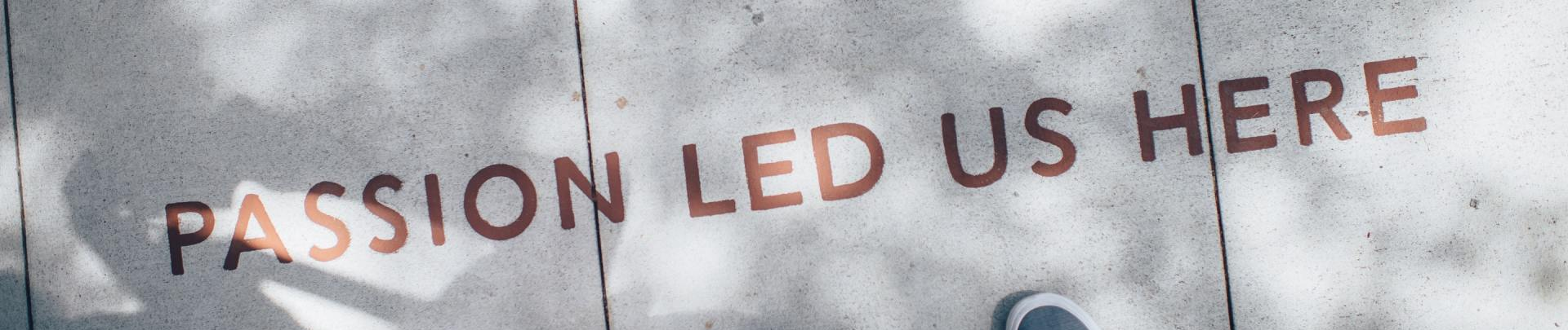 Two pairs of shoes on a sidewalk beside the words “PASSION LED US HERE” painted on the ground