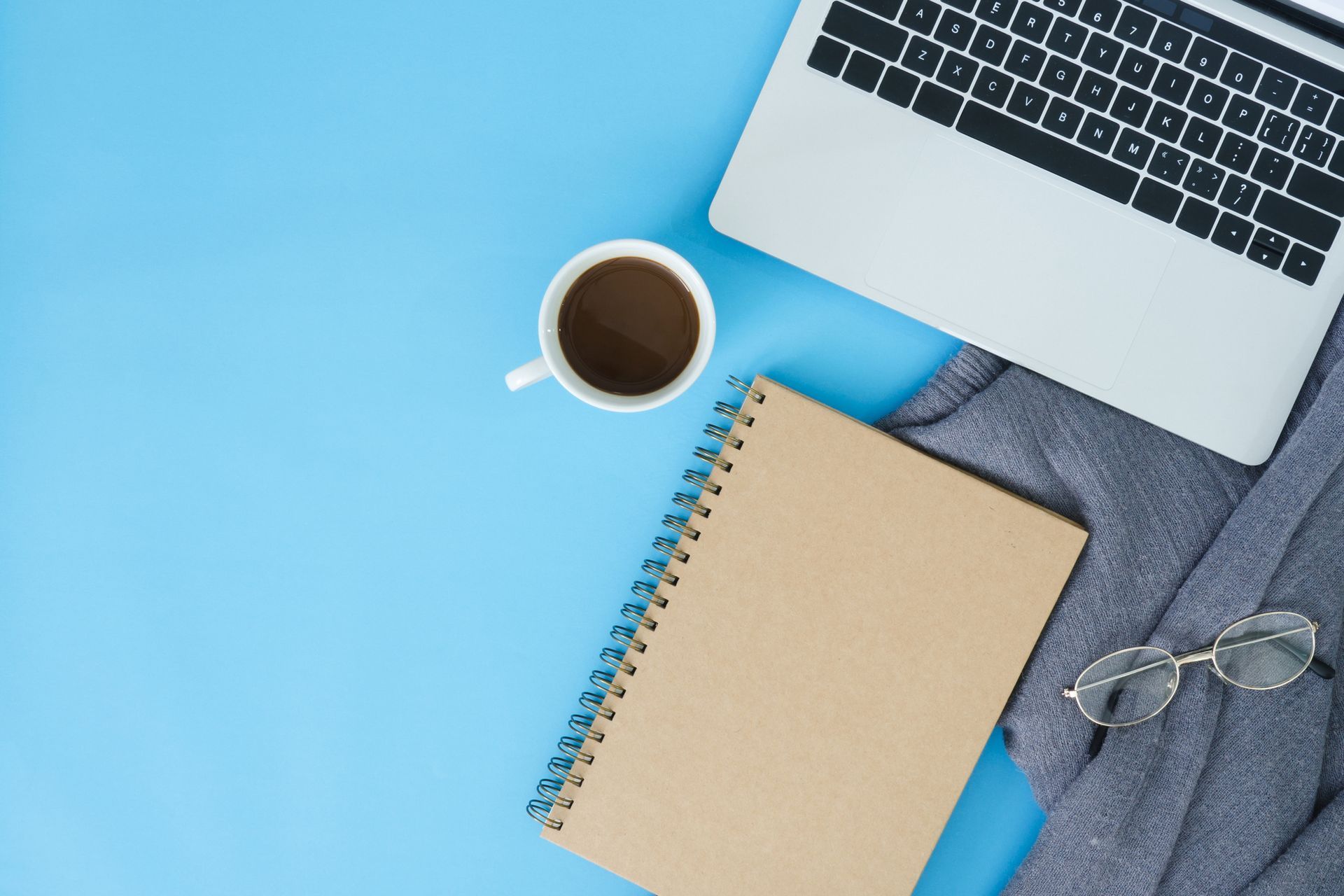 A laptop , notebook , cup of coffee and glasses on a blue table.
