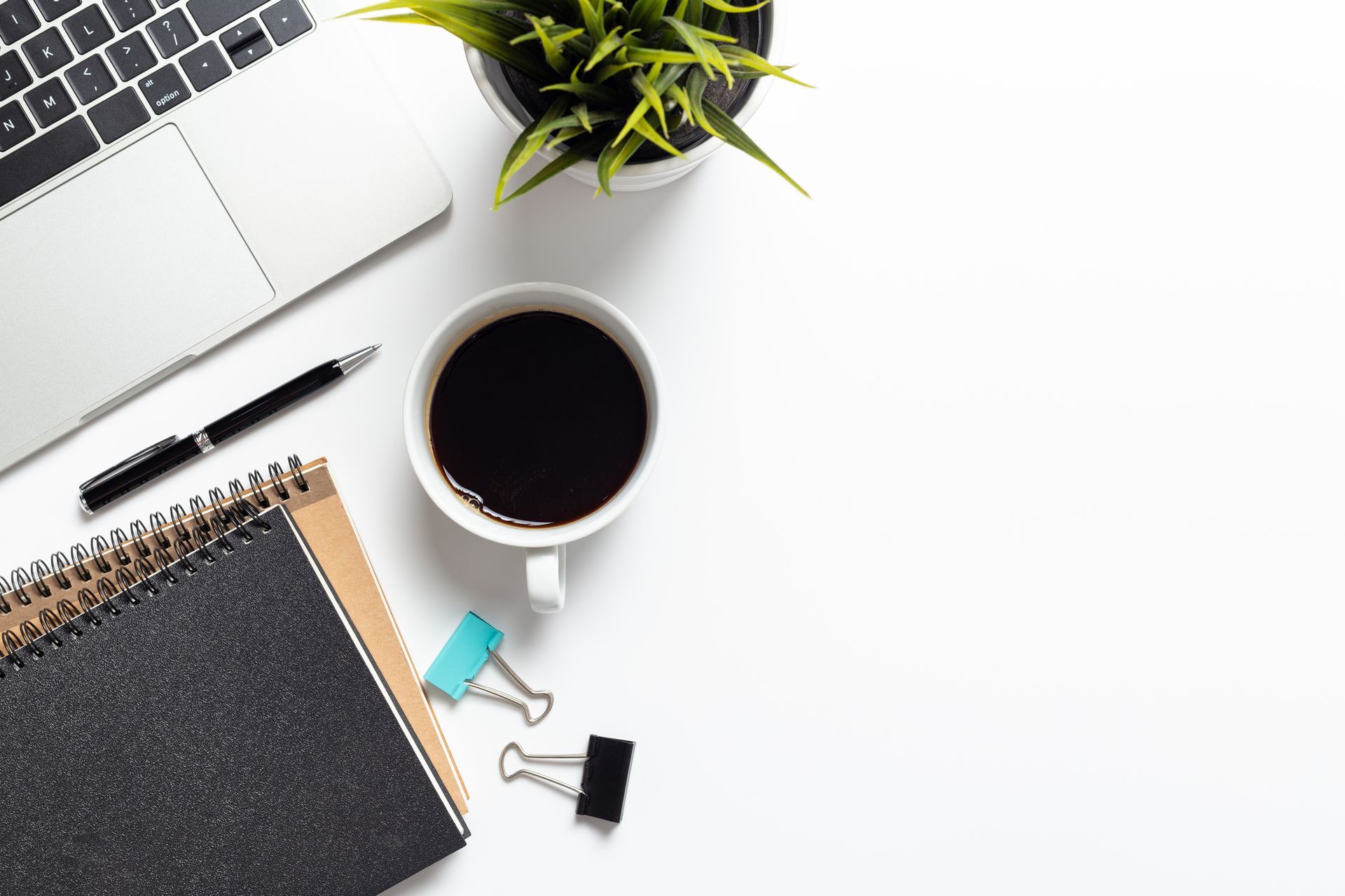 A cup of coffee , a notebook , a pen , and a laptop on a desk.