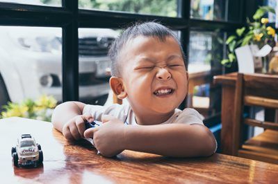Child smiling with eyes closed at a wooden table, holding a toy car near a bright window.