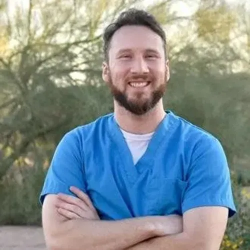 Smiling person in blue scrubs standing outdoors with arms crossed
