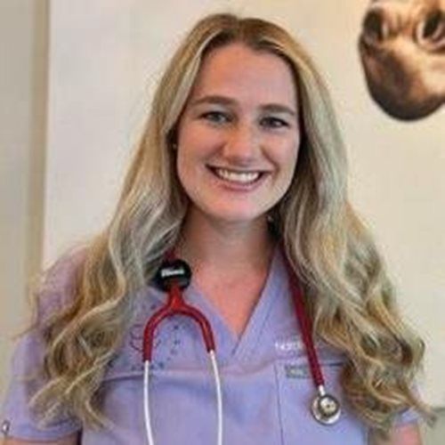 Smiling healthcare worker in a lavender scrubs top with a red stethoscope, standing indoors.