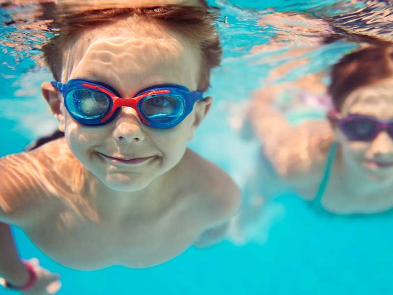 Two children underwater wearing goggles, smiling in a swimming pool