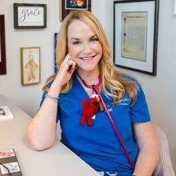 Woman smiling at a desk in a blue shirt with a red stethoscope, framed certificates on the wall.