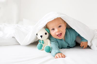 Baby in a teal sweater peeks out from under a white blanket beside a plush toy on a white bed