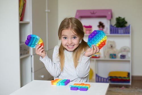 Smiling child holding rainbow pop-it toys at a white table in a playroom