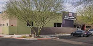 Low-rise beige building with a tree in front and a parked car beside a sign on the right.
