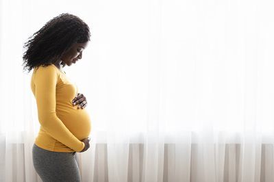 Pregnant woman in a yellow top standing in profile by a bright window, holding her belly