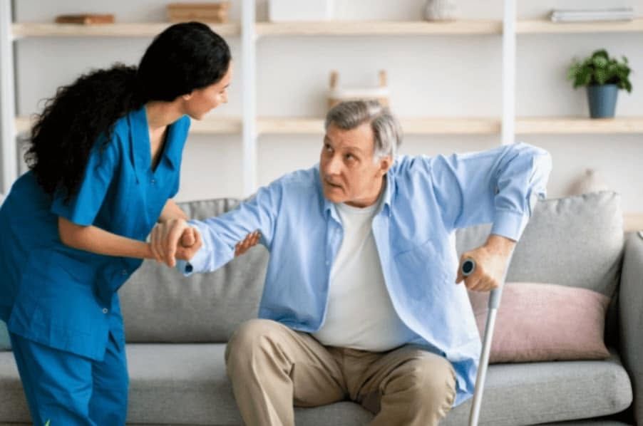 Caregiver helping a seated older adult up from a sofa in a bright living room
