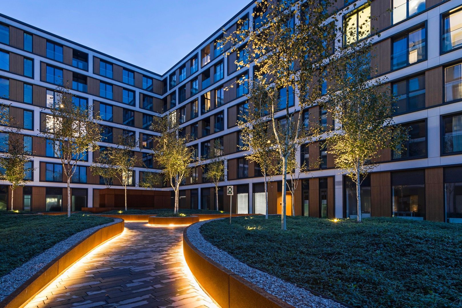 Cortile di un edificio moderno al tramonto, con alberi, sentieri illuminati e luci.