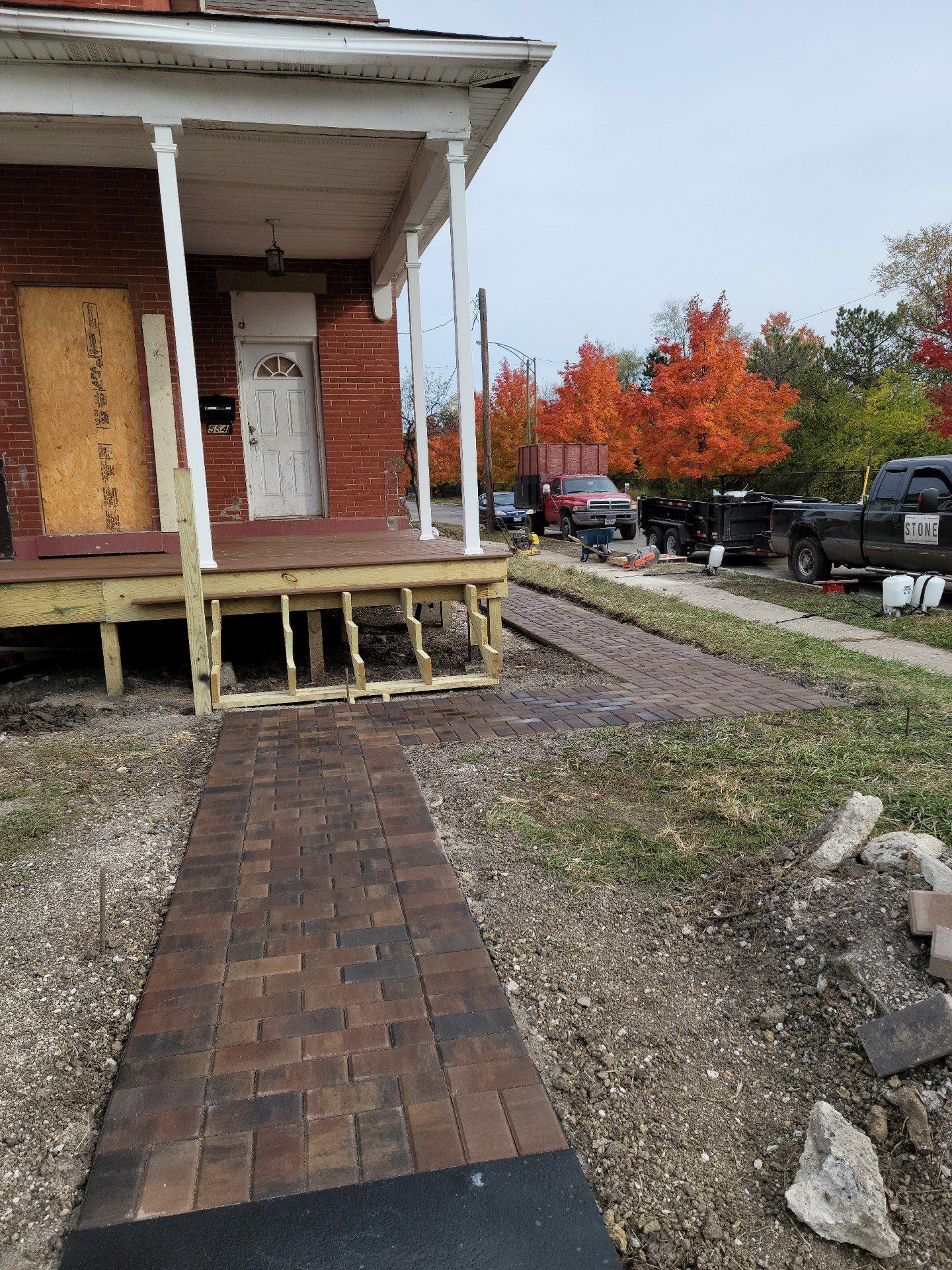 Brick walkway to an old porch with boarded windows, beside a gravel drive and autumn trees.