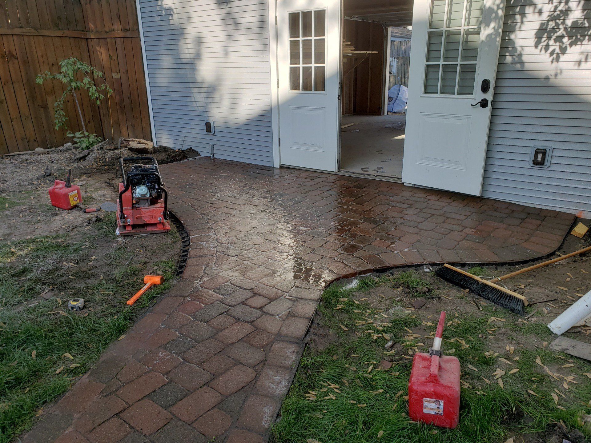 Brick walkway under construction outside open double doors, with tools and red gas cans on the ground.