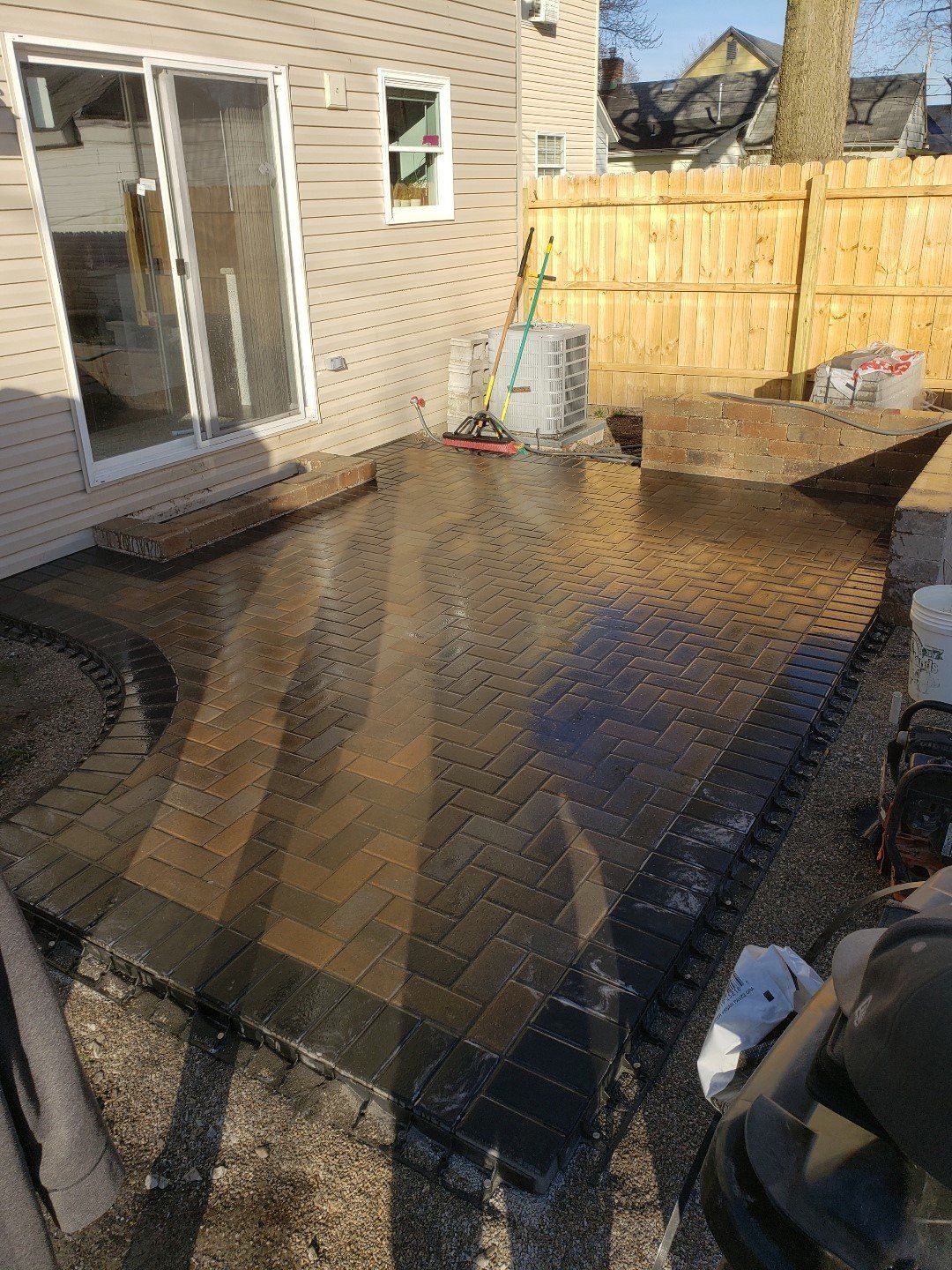 Backyard patio with a wet brick surface, sliding glass door, and wooden fence in sunlight