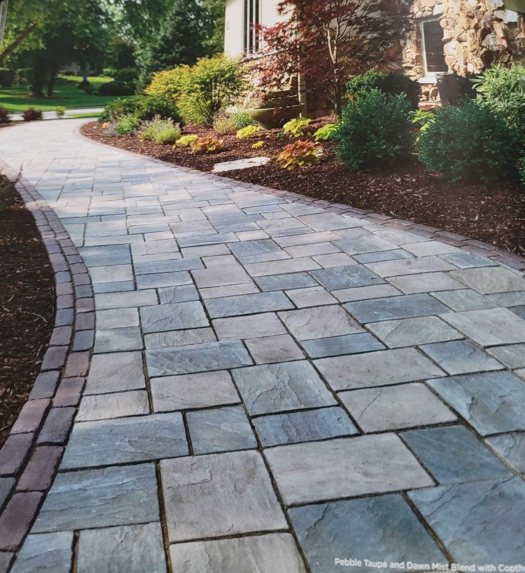 Curved stone paver walkway beside a landscaped house with shrubs and trees