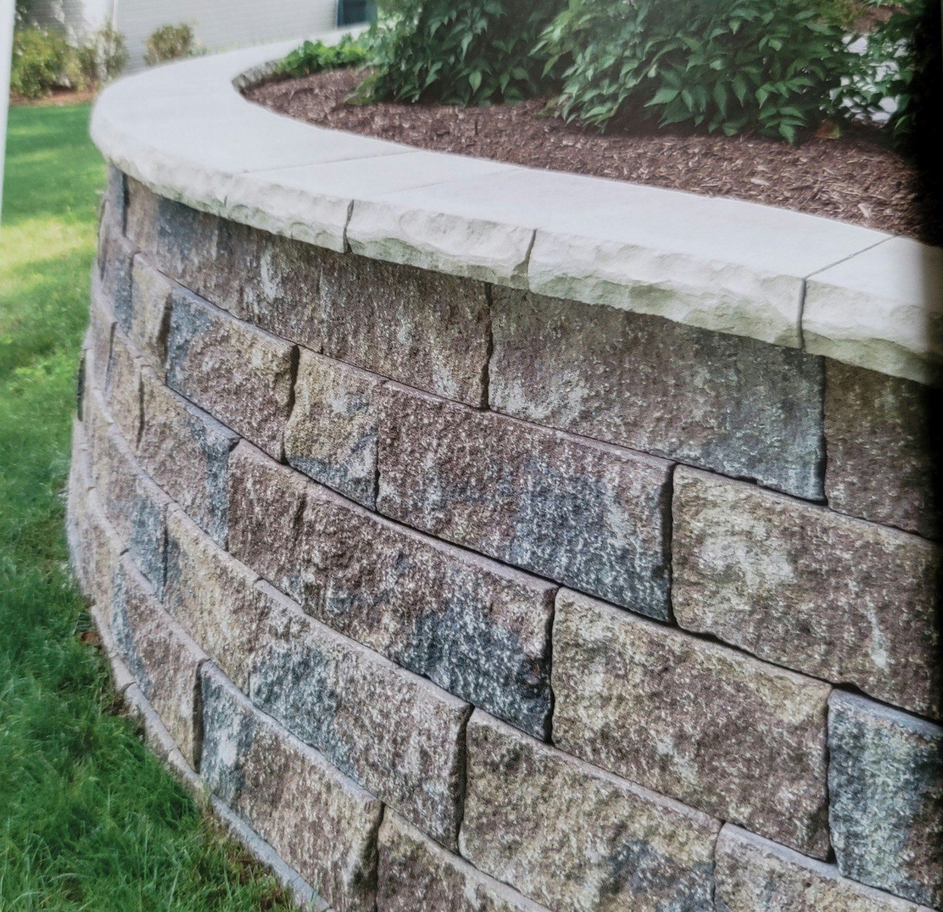 Curved stone retaining wall with a light cap beside green grass and shrubs outdoors