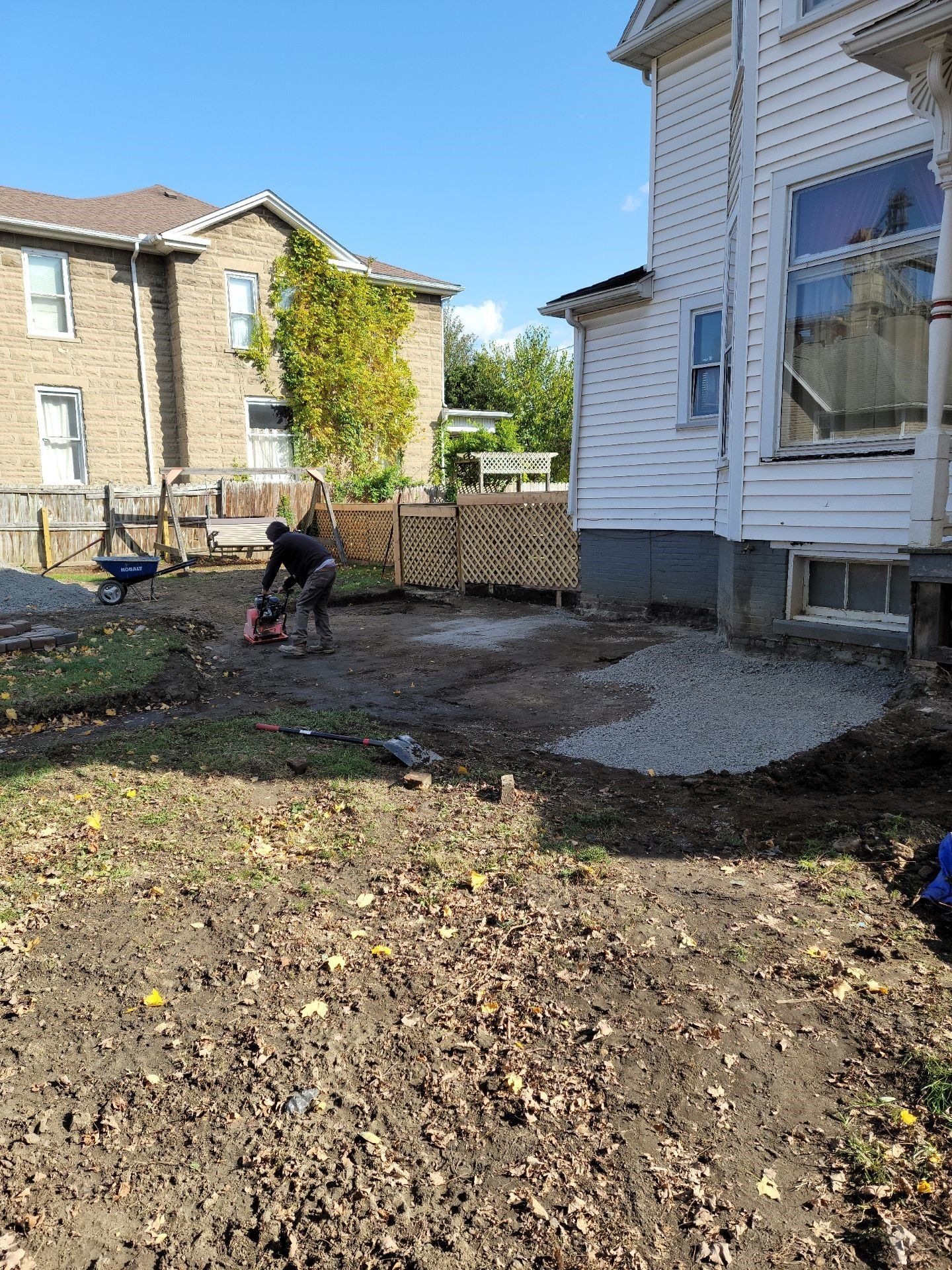 Backyard with gravel ground, a person working near a fence, and houses beside a white building.