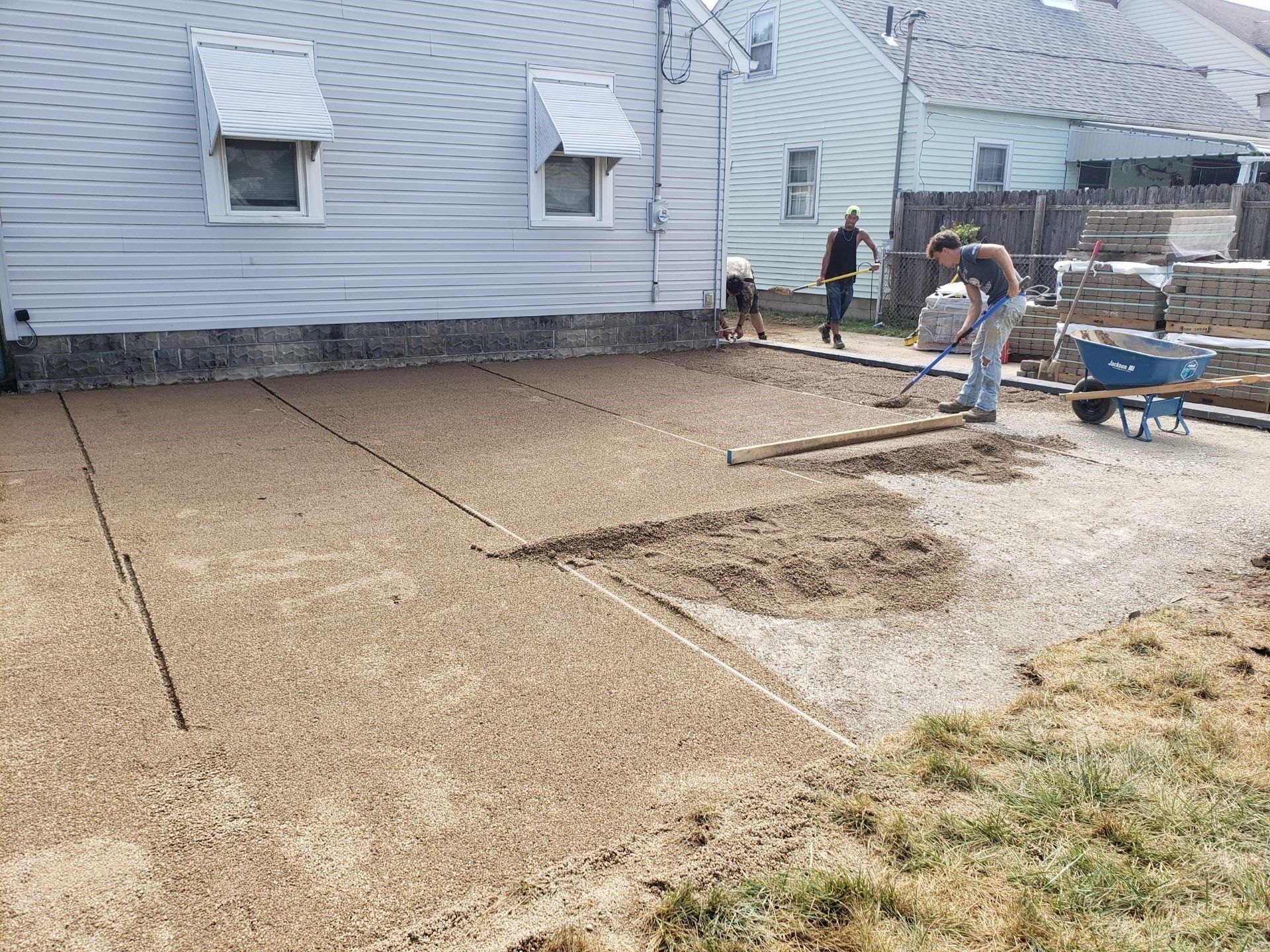 Workers smoothing concrete patio beside a gray house, with wheelbarrow and tools in a backyard.
