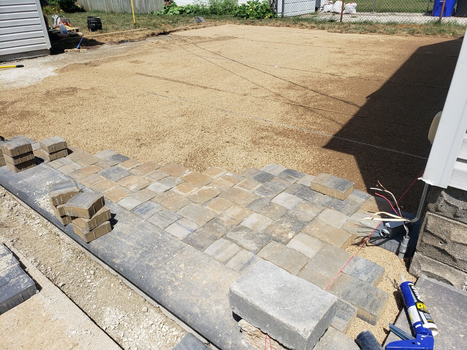 Partially paved gravel yard with bricks, concrete blocks, and tools near a building.