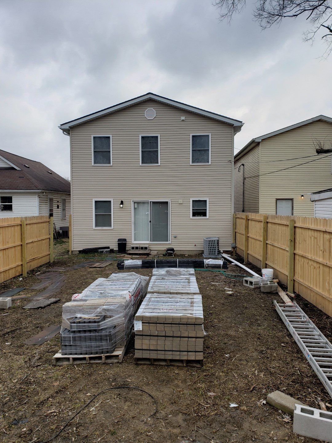 Backyard construction site with stacked pavers in front of a two-story house and fenced sides