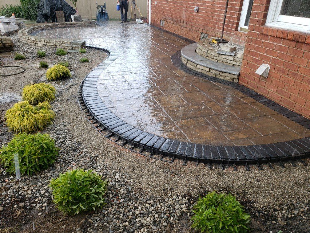 Curved brick patio and wet stone walkway beside a red brick house, bordered by gravel and small shrubs.