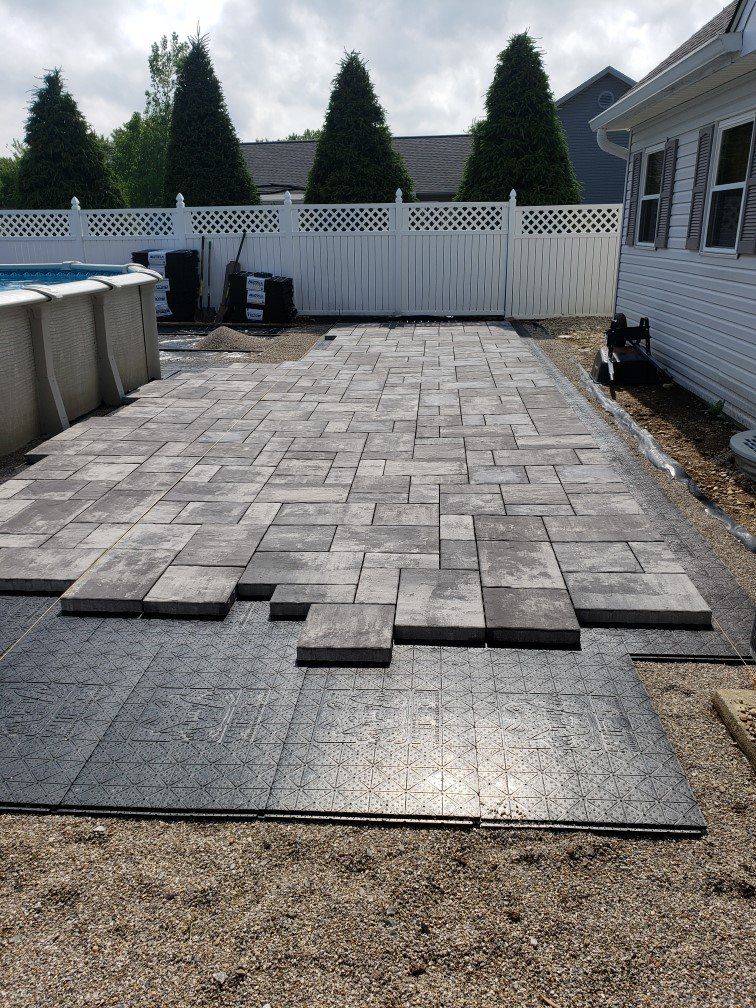 Partially installed gray paver patio beside a house, with loose pavers and gravel at the edge.