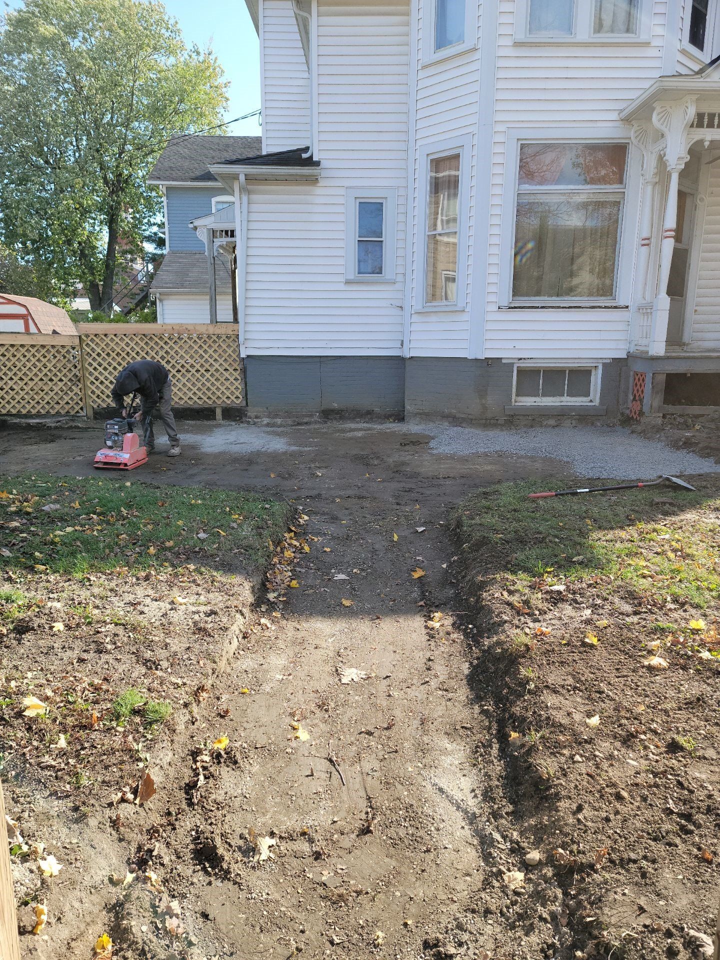 Worker edging a yard beside a white house, with a red tool on the ground.