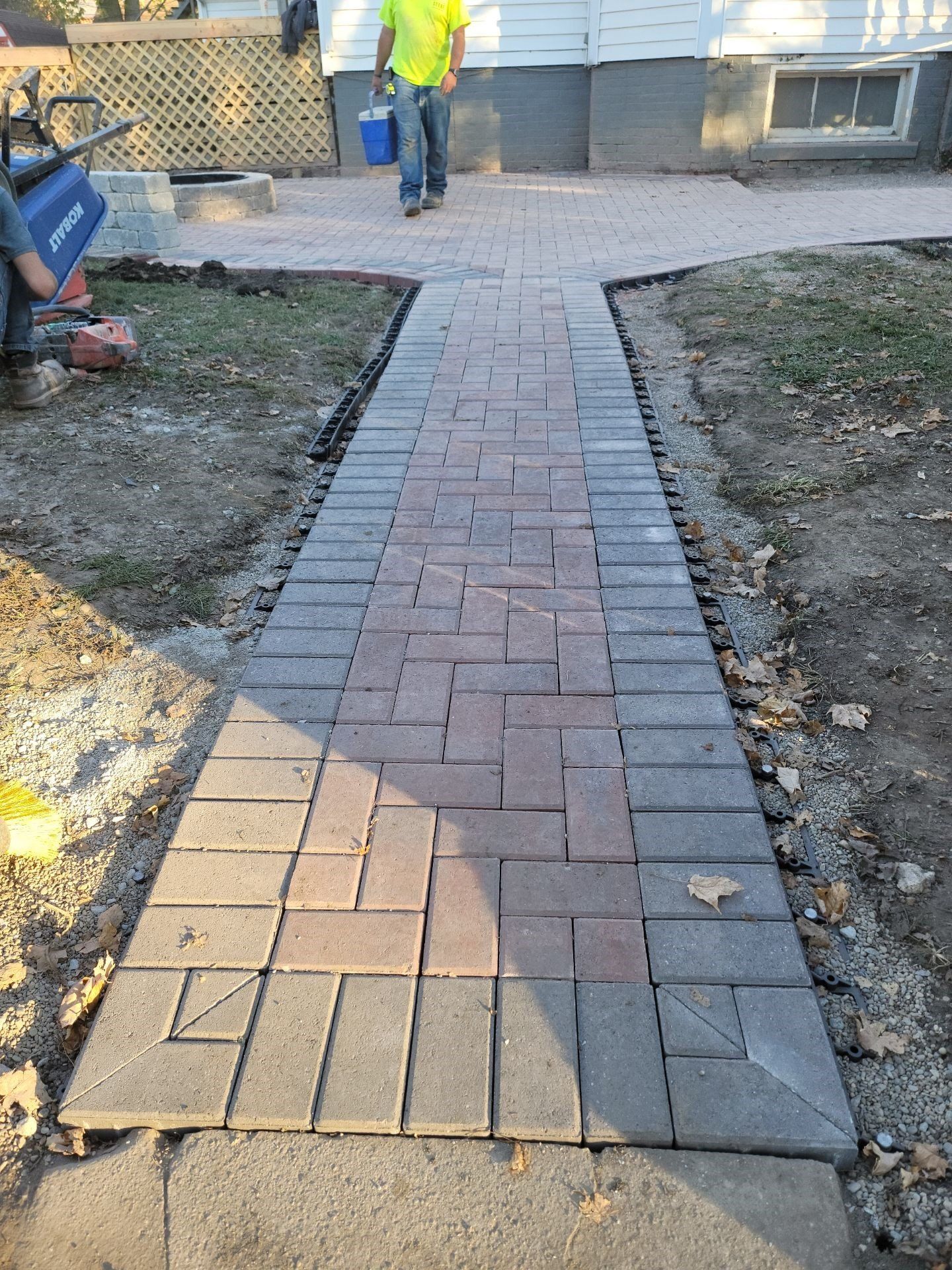 Brick walkway leading to a house entrance, bordered by dirt and grass with workers in the background