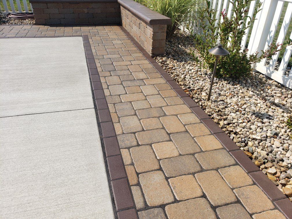 Paver walkway beside a concrete patio, brick wall, and gravel garden border with plants.