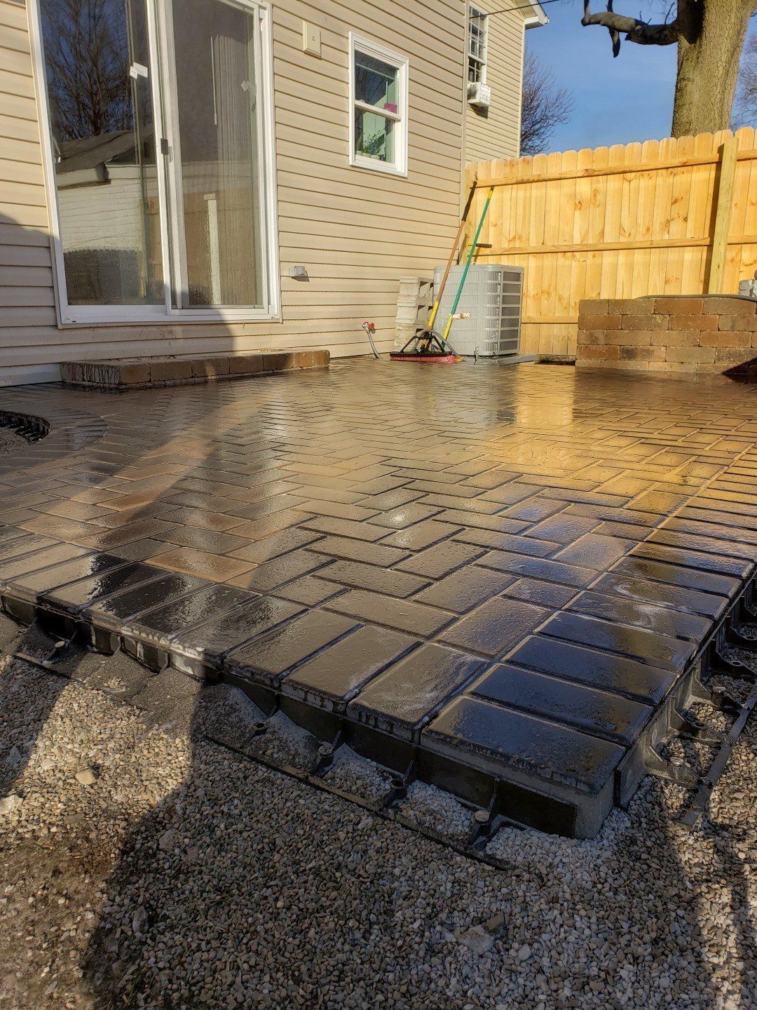 Wet brick patio beside a house and wooden fence, with a broom leaning against the wall.