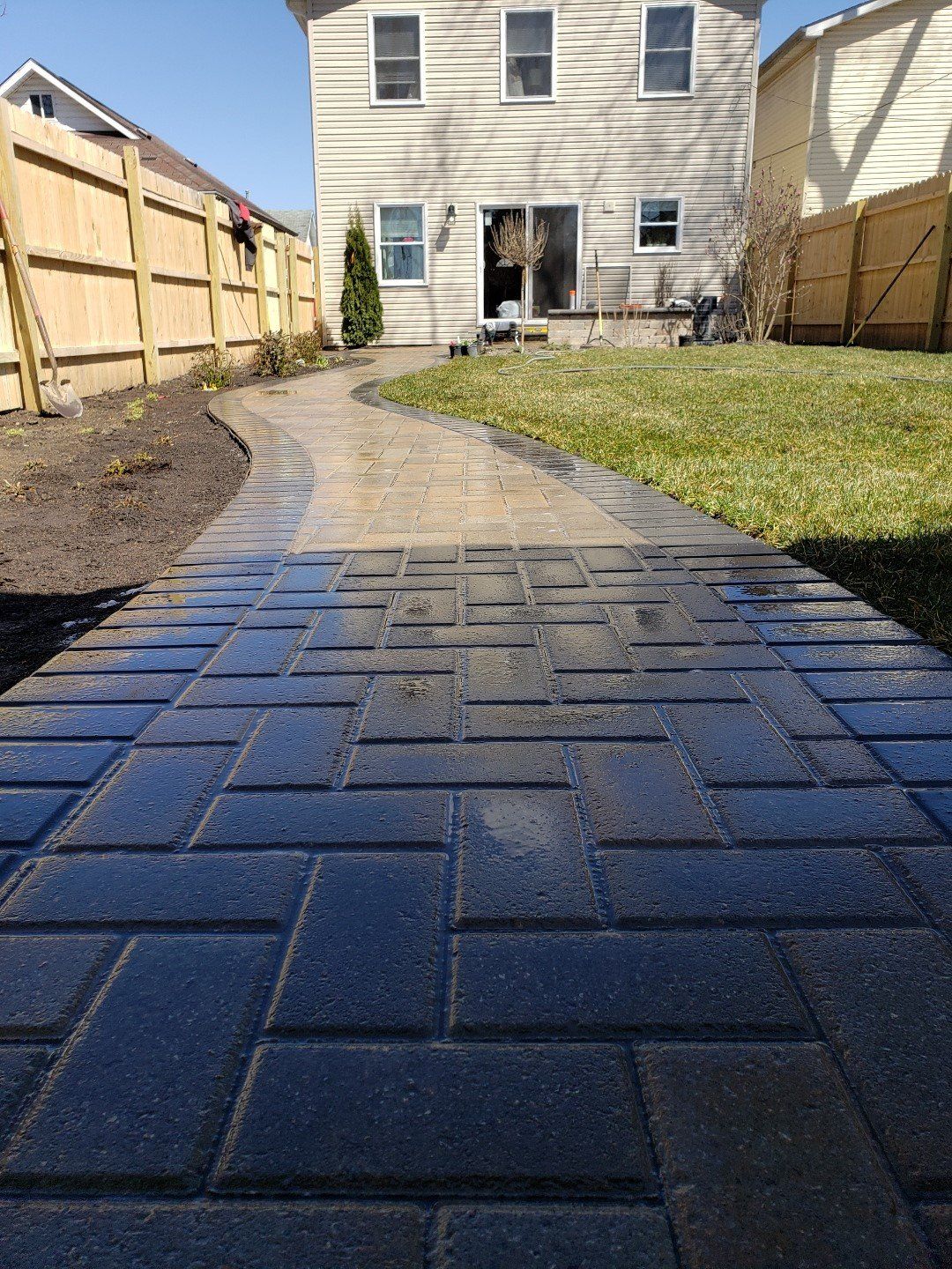 Black brick walkway leading to a house in a fenced backyard, with mulch beds on both sides.