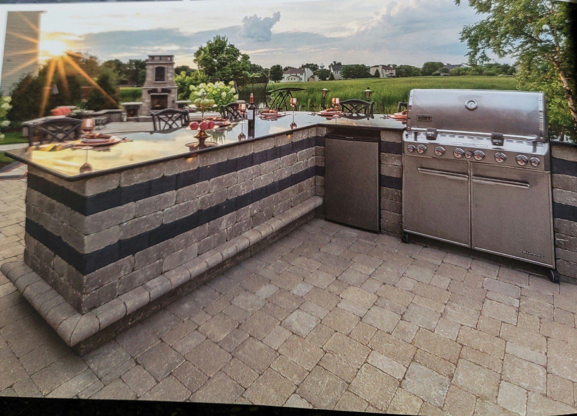 Outdoor patio with built-in grill, stone counter, and sunset view over green landscape.