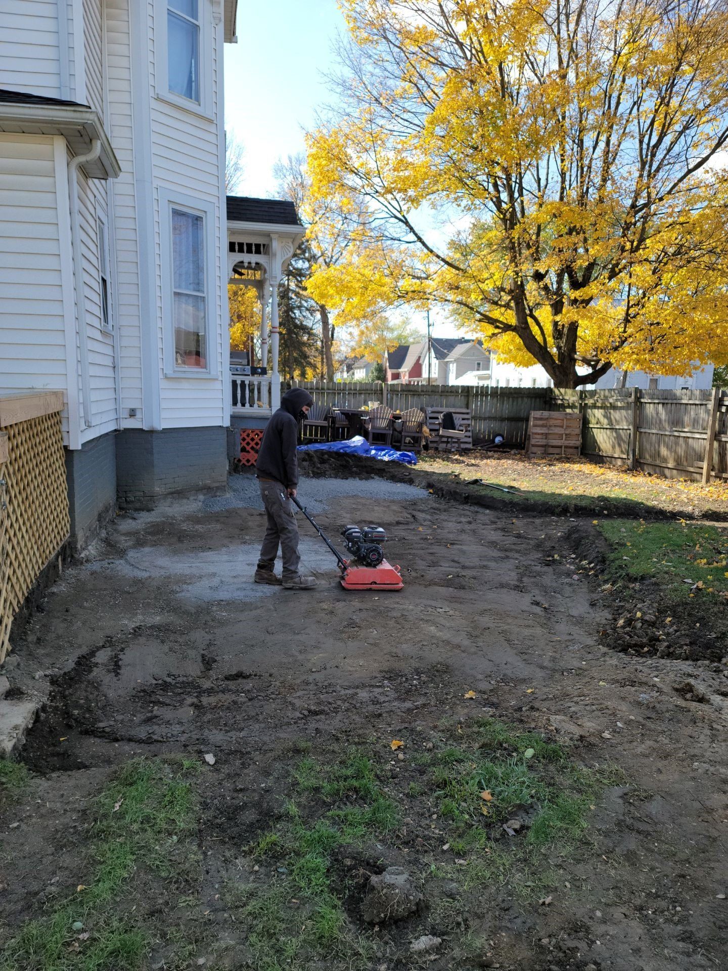 Person using a red lawn edger beside a house in a yard with autumn trees and a fence.